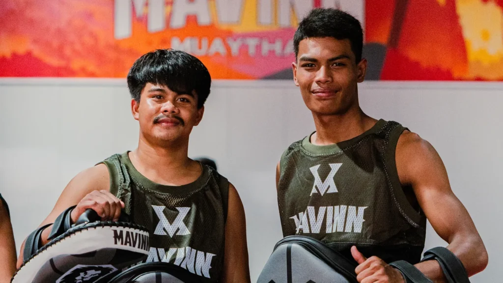 About Two students holding Thai pads in front of the MAVINN gym mural