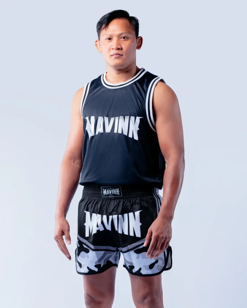 Home Kaonar P.K. Saenchai, Muay Thai trainer at MAVINN Muay Thai in Bangkok, standing in MAVINN team vest and camo Muay Thai shorts